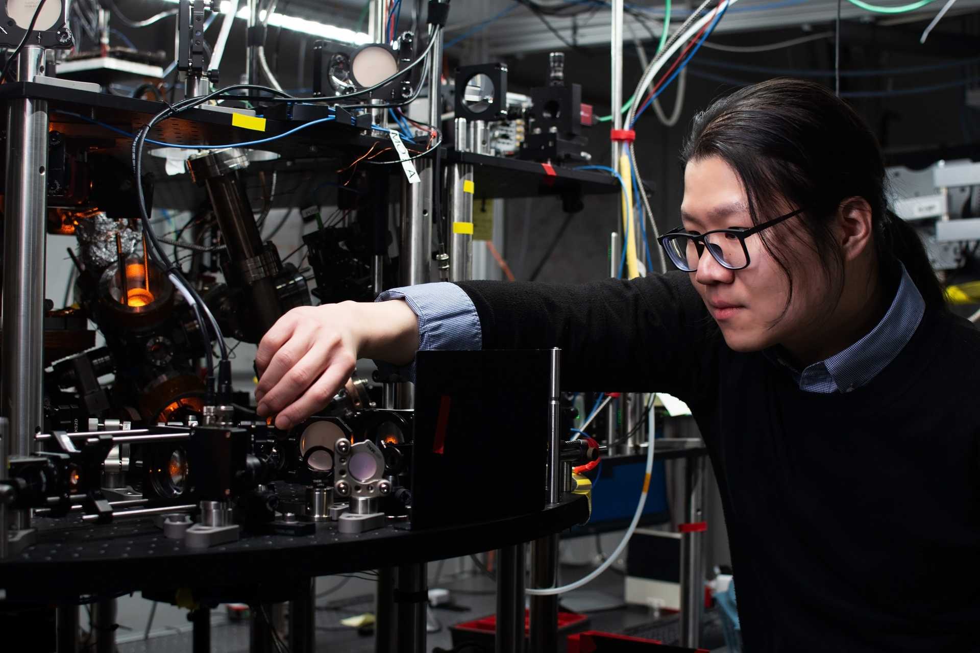 A man wearing glasses and a black sweater adjusts optical equipment in front of a cluttered lab space.
