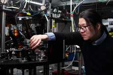 A man wearing glasses and a black sweater adjusts optical equipment in front of a cluttered lab space.